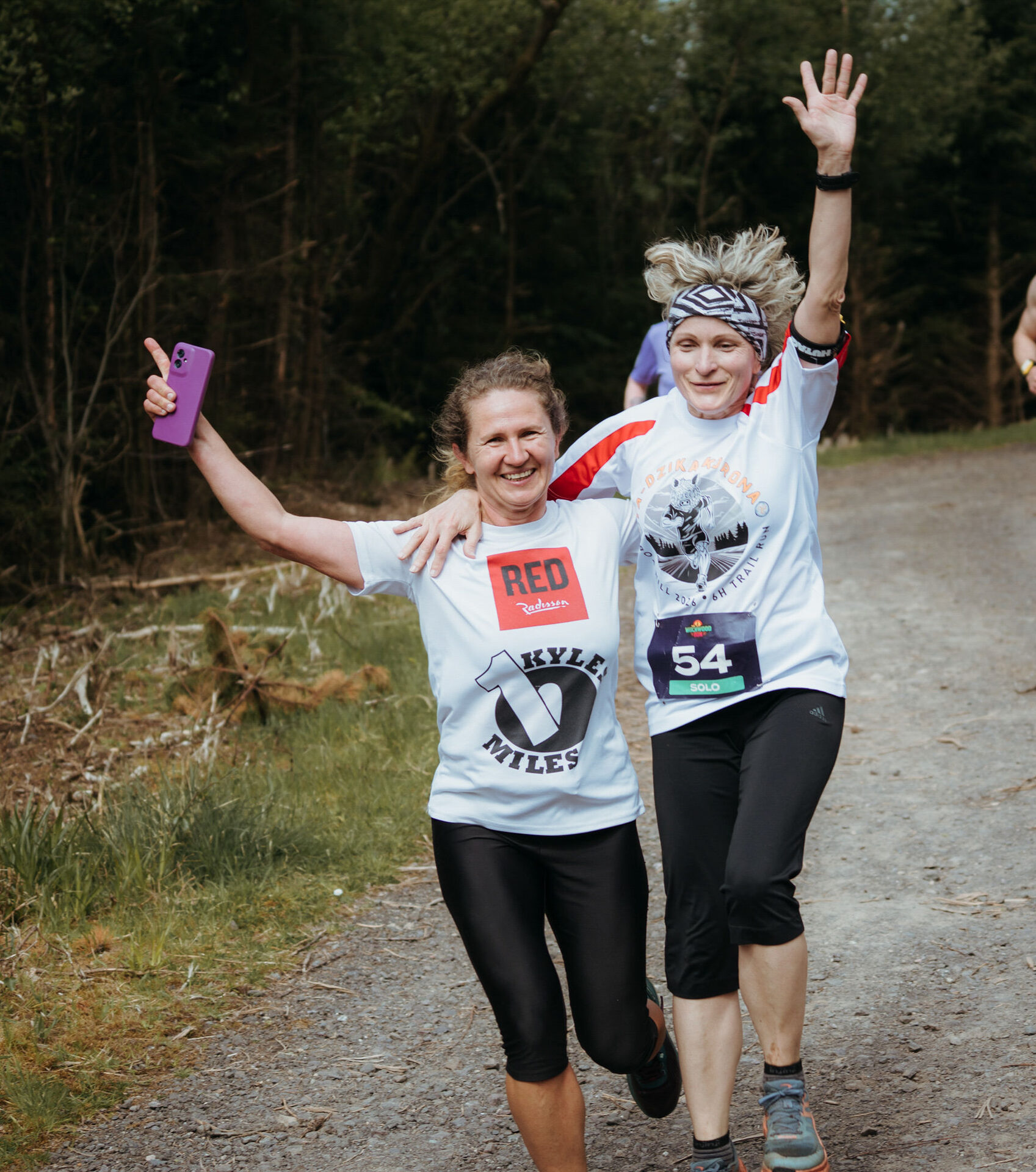 Two runners arm in arm, phone held high, celebrating on the trail