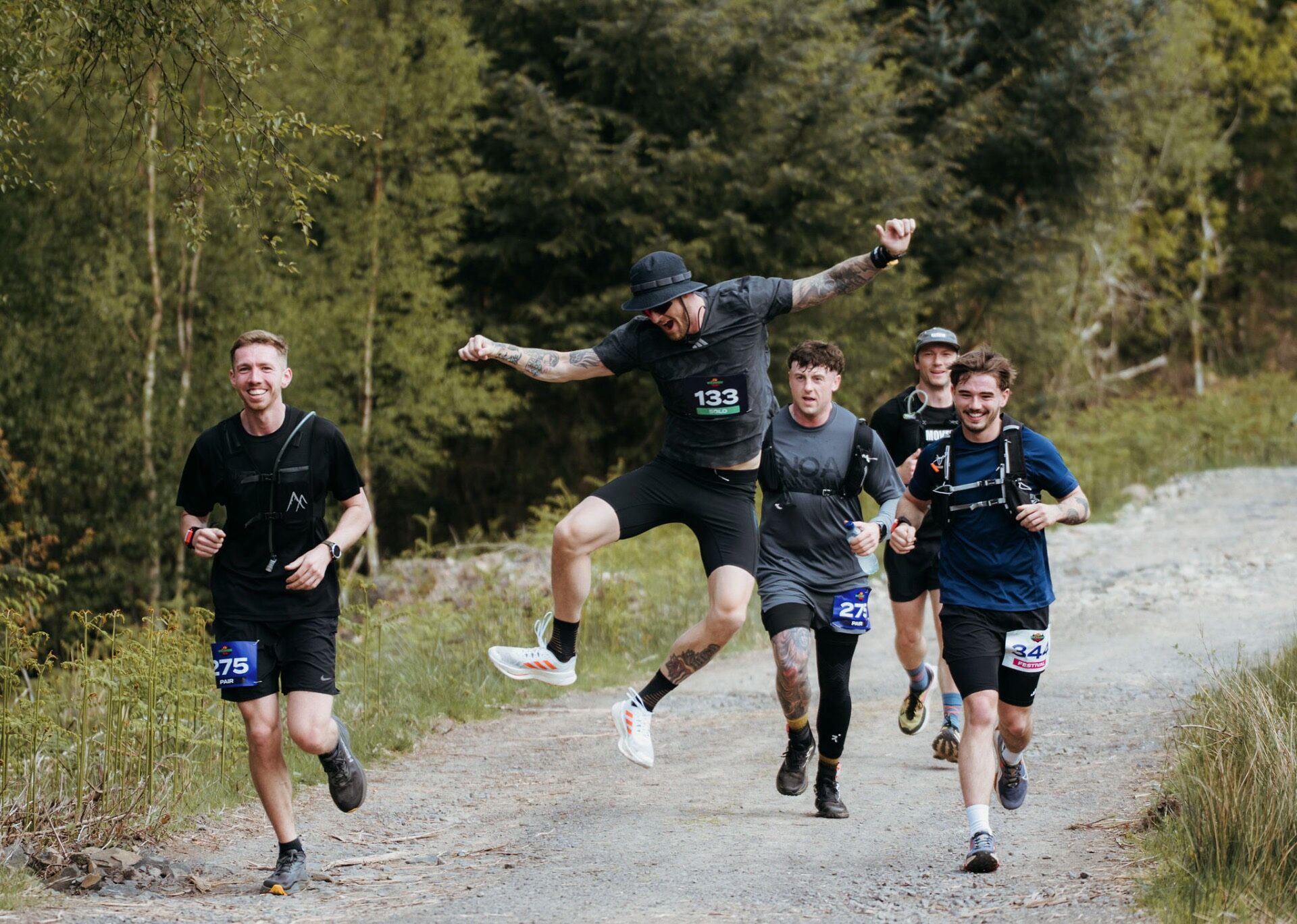Runner leaping mid-air with arms out, group cheering on the trail